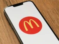 A smartphone lying on a wooden surface displays the McDonald’s yellow “M” logo inside a red circle on its screen. The phone is tilted slightly to the left, and the logo is centered on a white background.