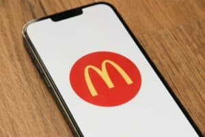 A smartphone lying on a wooden surface displays the McDonald’s yellow “M” logo inside a red circle on its screen. The phone is tilted slightly to the left, and the logo is centered on a white background.