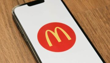 A smartphone lying on a wooden surface displays the McDonald’s yellow “M” logo inside a red circle on its screen. The phone is tilted slightly to the left, and the logo is centered on a white background.
