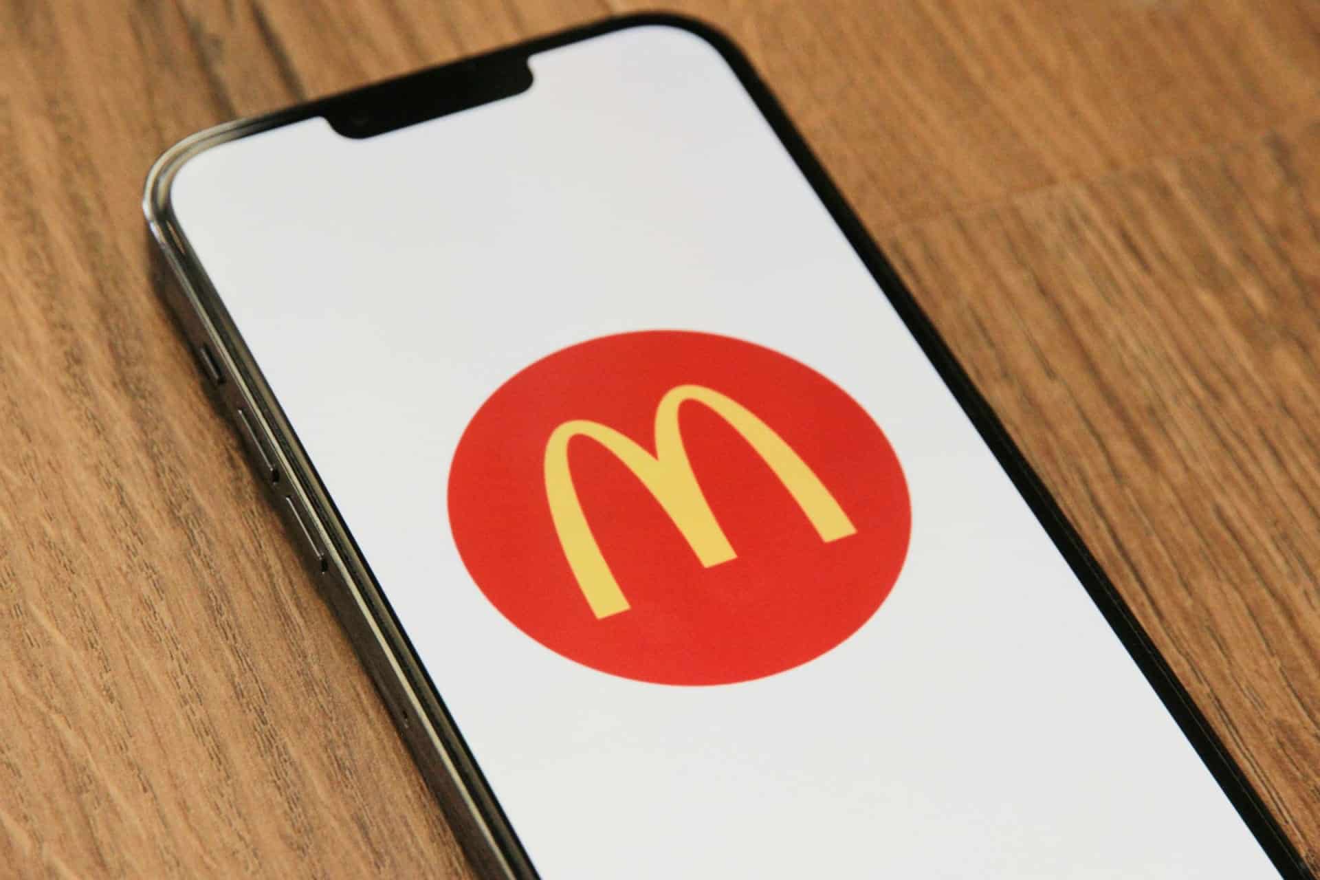 A smartphone lying on a wooden surface displays the McDonald’s yellow “M” logo inside a red circle on its screen. The phone is tilted slightly to the left, and the logo is centered on a white background.