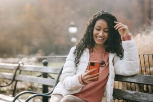 A woman with long curly hair is sitting on a bench, smiling at her phone. She wears a white jacket over a pink hoodie and has a fitness tracker on her wrist. The background shows a blurred park setting with autumn foliage, and the lighting is warm and soft.