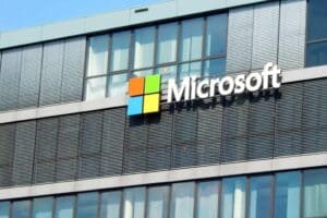 Glass building facade with the Microsoft logo. The logo features a square composed of four smaller colored squares: red, green, blue, and yellow, followed by the word "Microsoft" in white letters. The sky is clear and blue in the background.