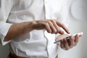 A person wearing a white long-sleeved shirt is holding a smartphone in their left hand and using their right index finger to touch the screen. The image focuses on the hands and the phone, with a blurred white background.