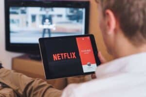 A person holding a tablet displaying the Netflix logo and text "11,521,823" on a screen split with a red section. In the background, a TV shows a blurred image of a person and text. The person is seated on a couch, wearing light-colored pants.