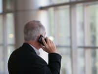 A man with gray hair wearing a dark suit is talking on a mobile phone. He is facing away, towards large windows with a blurred view of the outside. The setting appears to be indoors, possibly an office or airport terminal. The lighting is natural and soft.