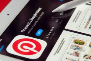 Close-up of an iPad screen displaying the Pinterest app page in the App Store. The page shows the "Pinterest: Lifestyle Ideas" app with a red logo and options to "GET" the app. It has a rating of 4.7 stars from 26K ratings. A stylus touches the screen near the app description.