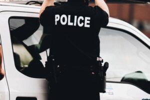 A person in a black shirt with "POLICE" in white text stands beside a white vehicle. The person has a utility belt with various equipment. The scene seems to be outdoors, possibly during the day, with a blurred background.