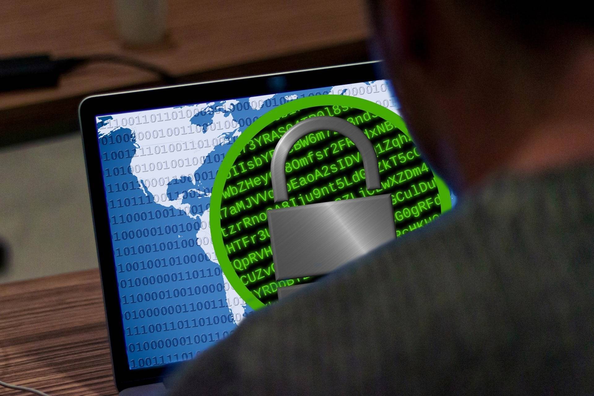 Person facing laptop displaying code in green text against a dark background, overlaid with a digital padlock symbol. The padlock is green with a gray shackle, indicating a theme of cybersecurity or data protection. A partial map is visible beneath the code.