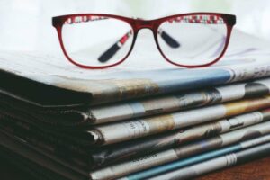 A pair of red-framed eyeglasses rests atop a stack of neatly folded newspapers. The newspapers are layered in a way that their edges create a pattern, with visible headlines and text. Soft, natural lighting filters in from the background, creating a calm and focused atmosphere.