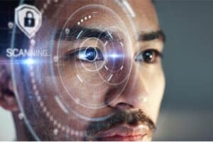 A close-up of a man's face focusing on his eye, surrounded by virtual holographic elements, suggesting an eye-scanning technology. A digital lock icon and the word "SCANNING..." appear on the left, indicating security or identity verification. The background is blurred.