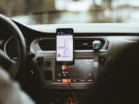 A smartphone displaying a navigation app is mounted on a car dashboard. The dashboard has a large display screen showing a radio channel, "RFM." The steering wheel and part of the driver's hand are partially visible. The background is blurred, indicating motion.
