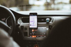 A smartphone displaying a navigation app is mounted on a car dashboard. The dashboard has a large display screen showing a radio channel, "RFM." The steering wheel and part of the driver's hand are partially visible. The background is blurred, indicating motion.