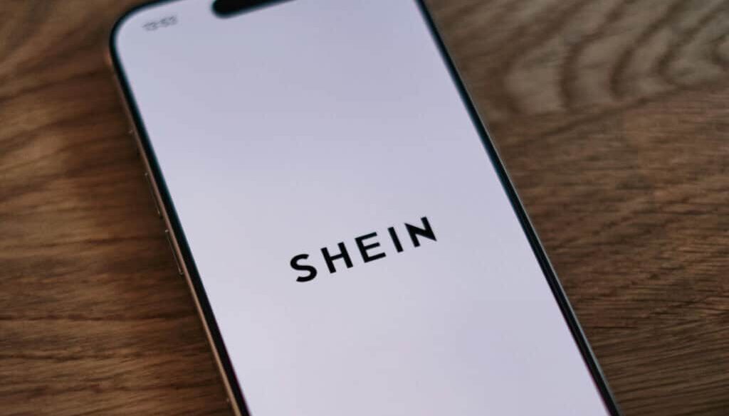 A smartphone with a white screen displays the word "SHEIN" in bold, black letters. The phone is placed on a wooden surface, viewed from a slight angle.