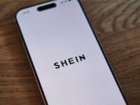 A smartphone with a white screen displays the word "SHEIN" in bold, black letters. The phone is placed on a wooden surface, viewed from a slight angle.