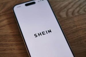 A smartphone with a white screen displays the word "SHEIN" in bold, black letters. The phone is placed on a wooden surface, viewed from a slight angle.