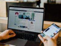 A person is using a laptop showing a Facebook profile while holding a smartphone. The laptop screen displays a user's cover and profile photo, with posts beneath. The person sits at a wooden desk in a bright room. The smartphone's screen shows a blurred app interface.