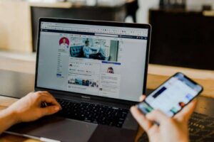 A person is using a laptop showing a Facebook profile while holding a smartphone. The laptop screen displays a user's cover and profile photo, with posts beneath. The person sits at a wooden desk in a bright room. The smartphone's screen shows a blurred app interface.