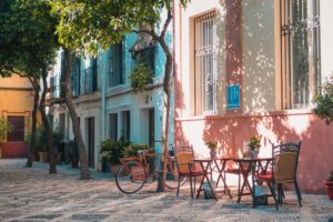 A charming, sunlit courtyard features a cobblestone path lined with trees. On the right, a set of outdoor tables with chairs sits against colorful building facades in pastel shades. A vintage red bicycle is parked nearby, and potted plants accentuate the tranquil scene.