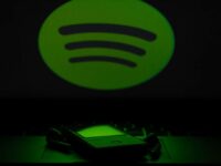 A dark image features a large green Spotify logo on a black background. In the foreground, an iPhone and a pair of earbuds rest on a reflective surface. The scene is predominantly green, with the light from the logo casting a subtle glow on the phone and earbuds.