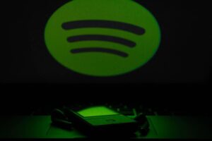 A dark image features a large green Spotify logo on a black background. In the foreground, an iPhone and a pair of earbuds rest on a reflective surface. The scene is predominantly green, with the light from the logo casting a subtle glow on the phone and earbuds.