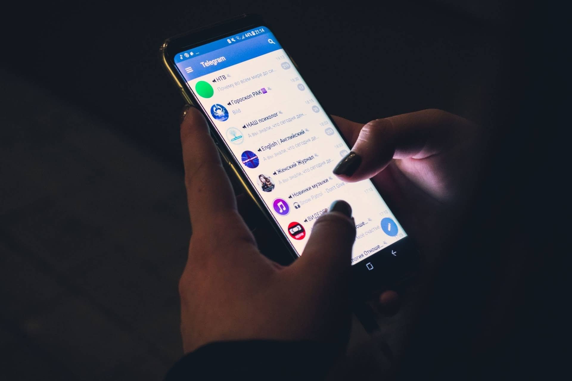 A person holds a smartphone displaying a chat list on the Telegram app. The screen is filled with various conversations, each with an icon and a brief message preview. The scene is dimly lit, highlighting the phone and hands, with focus on the app's interface.