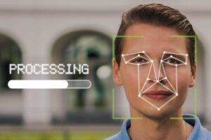 A man with short hair and a slight smile is facing forward. There is a digital overlay on his face displaying facial recognition features, with lines and points marking facial geometry. To the left, the word "PROCESSING" is shown above a progress bar. The background is blurred.