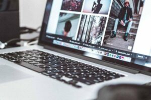 Open laptop displaying a series of photographs, featuring portraits and outdoor scenes. The keyboard is visible, and the background is slightly blurred with some equipment suggesting an office or studio setting. The screen shows images in a grid layout.