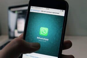 Close-up of a hand holding a smartphone displaying the WhatsApp app on the screen. The screen shows the WhatsApp logo, a green speech bubble with a white phone icon, over a green patterned background. The text below the logo reads "WhatsApp Version 2.12.13".
